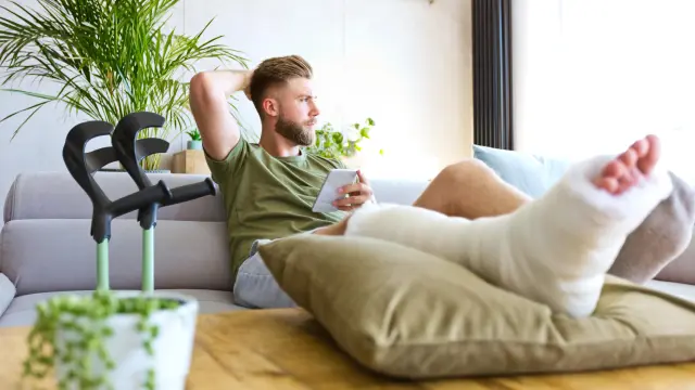 A man with a beard is sitting on a grey couch, his legs are on the table in front of him. His right leg is in a cast up to the knee. He is looking out the window, holding his mobile phone in his left hand, his crutches are next to him.