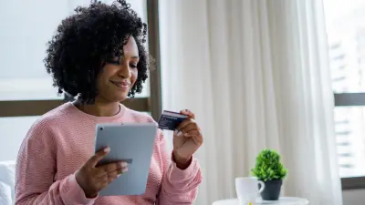 A woman with brown curly hair wearing a pink sweater sits on the couch and holds her health insurance card in her left hand. In the other hand she holds a tablet.