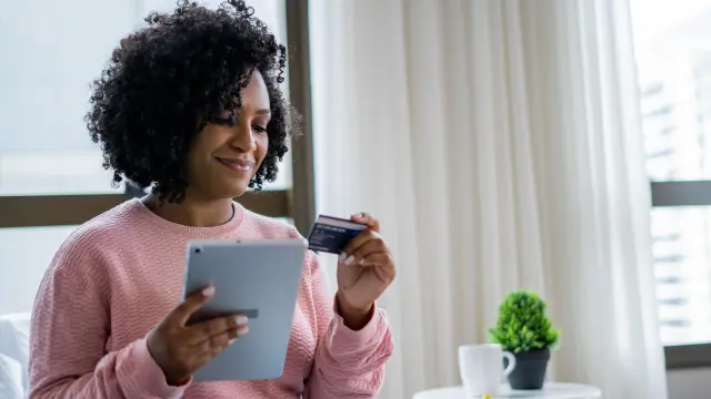 A woman with brown curly hair wearing a pink sweater sits on the couch and holds her health insurance card in her left hand. In the other hand she holds a tablet.