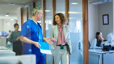 A sales representative and a doctor are walking side by side in an office corridor, other employees can be seen in the background.