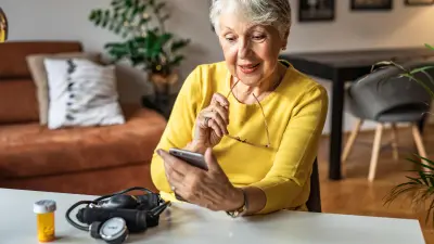 An elderly lady with gray hair is holding her glasses, sitting at the table in her living room and looking at her smartphone. On the table is a sphygmomanometer and a tube of pills.