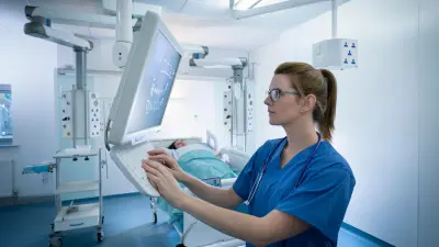 A doctor is wearing a blue surgical shirt and is using the keyboard of a computer. A patient can be seen in a bed in the background.