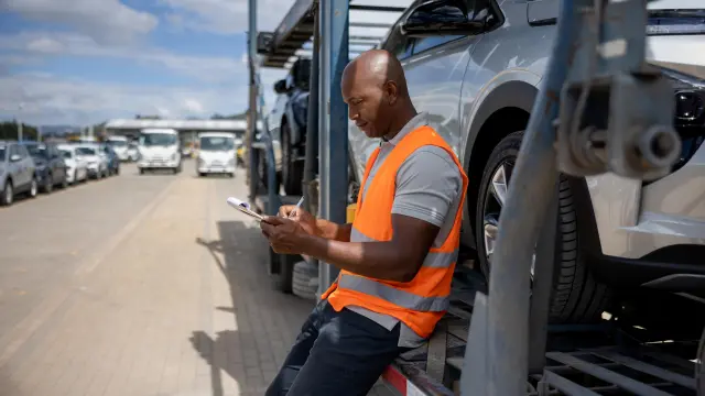 A man leans against the trailer of his truck, writing something on a clipboard. In the background, a blur of various vehicles can be seen.